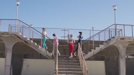 Grupo-Multiétnico-De-Mujeres-Corriendo-Sobre-Las-Escaleras-De-Un-Puente-En-La-Playa-Y-El-Fondo-Del-Cielo-Azul