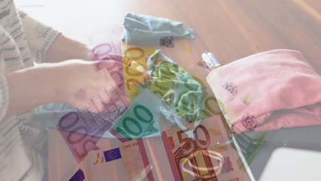 Digital-composite-video-of-Euro-bills-moving-against-woman-sewing-face-mask-in-background