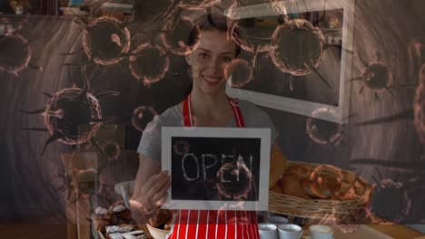 Animation-of-macro-Covid-19-cells-floating-over-happy-Caucasian-female-chef-holding-Open-sign