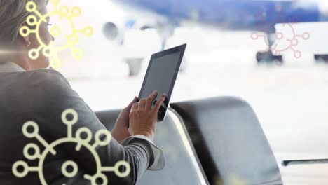 Animation-of-floating-Covid-19-cell-icons-over-Caucasian-woman-using-tablet-at-airport