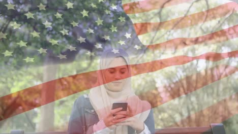 Animación-De-La-Bandera-Estadounidense-Ondeando-Sobre-Una-Mujer-De-Raza-Mixta-Con-Hijab-Usando-Un-Teléfono-Inteligente.