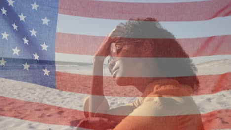 Animation-of-American-flag-waving-over-woman-with-sunglasses-sitting-on-beach-by-seaside