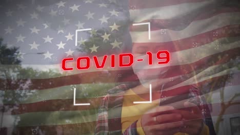Texto-Covid-19-Y-Bandera-Estadounidense-Ondeando-Contra-Una-Mujer-Que-Usa-Mascarilla-Usando-Un-Teléfono-Inteligente