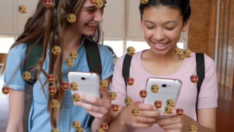 Multiple-face-emojis-floating-against-two-female-students-using-smartphones