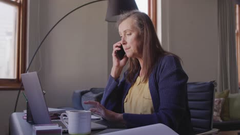 Mujer-Hablando-Por-Teléfono-Inteligente-Mientras-Usa-Una-Computadora-Portátil-En-Casa