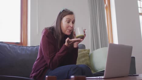 Woman-holding-coffee-cup-having-a-video-chat-on-her-laptop-at-home