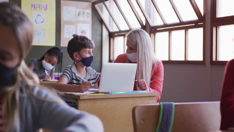 Female-teacher-and-boy-wearing-face-mask-using-laptop-in-class-at-school