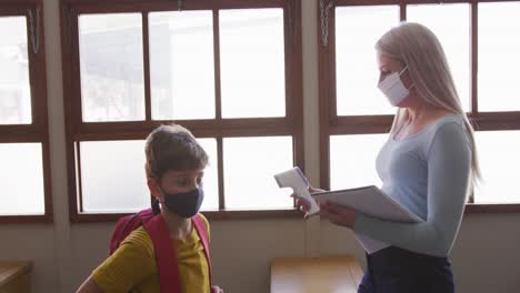 Female-teacher-wearing-a-face-mask-measuring-temperature-of-a-boy-in-class-at-school