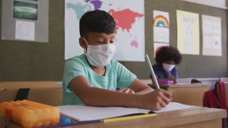 Junge-Mit-Gesichtsmaske-Schreibt,-Während-Er-In-Der-Schule-Auf-Seinem-Schreibtisch-Sitzt