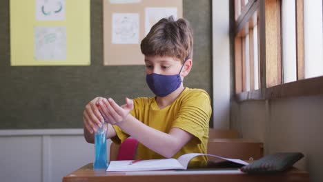 Junge-Mit-Gesichtsmaske-Desinfiziert-Seine-Hände,-Während-Er-In-Der-Schule-Auf-Seinem-Schreibtisch-Sitzt