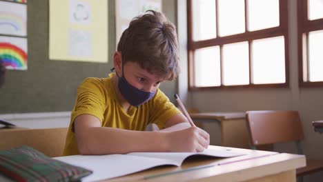Junge-Mit-Gesichtsmaske-Schreibt,-Während-Er-In-Der-Schule-Auf-Seinem-Schreibtisch-Sitzt