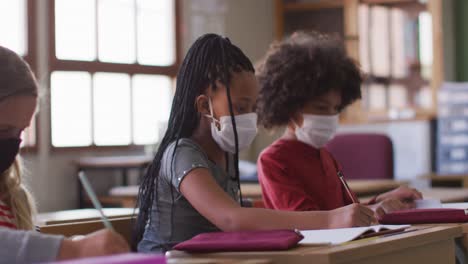 Group-of-kids-wearing-face-masks-writing-while-sitting-on-their-desk-at-school