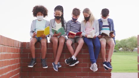 Gruppe-Von-Kindern-Mit-Gesichtsmasken,-Die-Bücher-Lesen,-Während-Sie-Auf-Einer-Ziegelwand-Sitzen