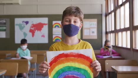 Junge-Trägt-Gesichtsmaske-Und-Hält-Ein-Regenbogengemälde-Im-Unterricht-In-Der-Schule