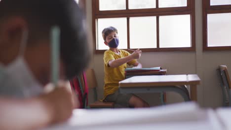 Junge-Mit-Gesichtsmaske-Niest,-Während-Er-In-Der-Schule-Auf-Seinem-Schreibtisch-Sitzt