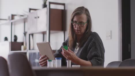 Mujer-Sosteniendo-Un-Contenedor-De-Medicamentos-Y-Usando-Una-Tableta-Digital-En-Casa