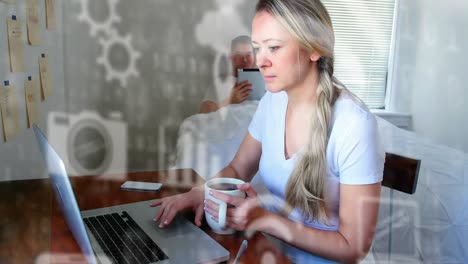 Multiple-digital-icons-floating-against-woman-with-coffee-cup-using-laptop-at-home