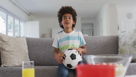 African-American-boy-at-home-sitting-on-a-sofa-in-the-living-room,-watching-sports-on-TV,-holding-a-