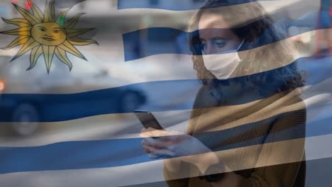 Bandera-Uruguaya-Ondeando-Contra-Una-Mujer-Con-Mascarilla-Usando-Un-Teléfono-Inteligente