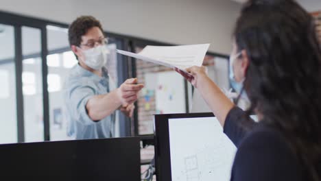 Man-wearing-face-mask-passing-a-document-to-his-colleague-at-office