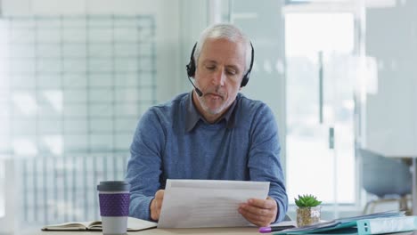 Man-wearing-headset-talking-while-sitting-on-his-desk-at-office