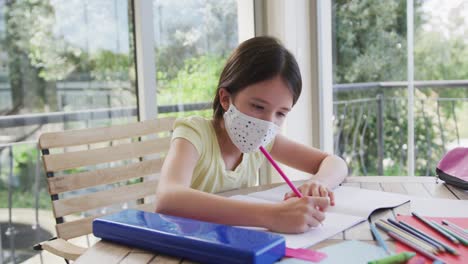 Caucasian-girl-spending-time-at-home-wearing-face-mask,-sitting-at-table-doing-her-homework,-in-slow