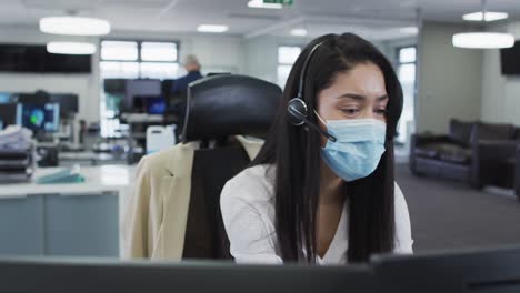 Woman-wearing-face-mask-talking-using-headset-at-office