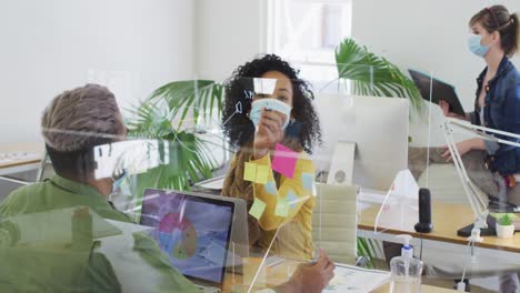 Woman-wearing-face-mask-writing-on-glass-board-on-her-desk-at-office