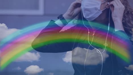 Rainbow-and-blue-sky-against-woman-wearing-face-mask-listening-to-music