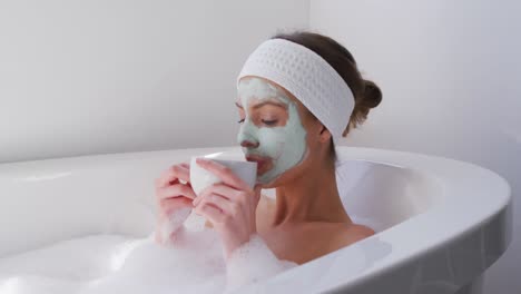 Frau-Trägt-Gesichtsmaske-Und-Trinkt-Kaffee,-Während-Sie-In-Der-Badewanne-Entspannt