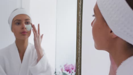 Frau-Im-Bademantel-Trägt-Gesichtsmaske-Auf,-Während-Sie-In-Den-Spiegel-Schaut