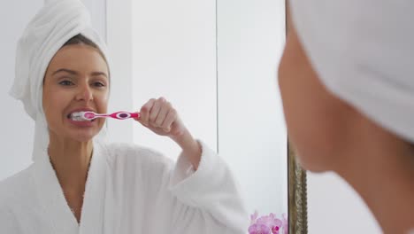 Woman-in-bathrobe-brushing-teeth-while-looking-in-the-mirror