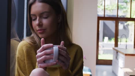 Woman-with-coffee-cup-looking-out-of-window-at-home