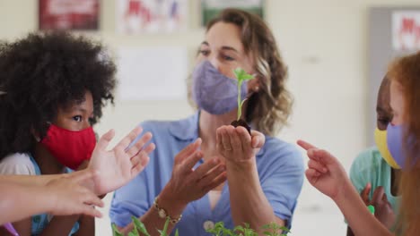 Female-teacher-wearing-face-mask-showing-plant-sapling-to-students-in-class