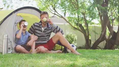 Caucasian-man-sitting-by-a-tent-with-his-son-in-the-garden-drinking-cups-of-tea