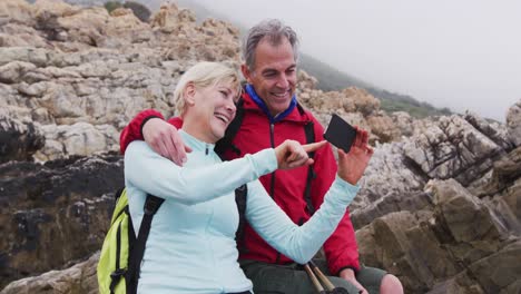 Senior-hiker-couple-with-backpacks-sitting-on-the-rocks-and-smiling-while-having-a-video-call