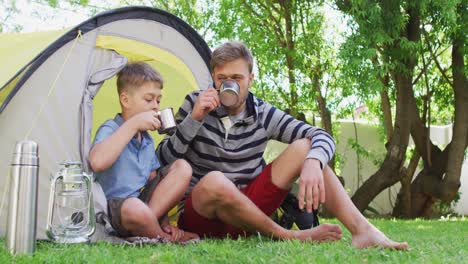 Caucasian-man-sitting-by-a-tent-with-his-son-in-the-garden-drinking-cups-of-tea-and-laughing-in-slow