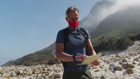 Senior-hiker-man-wearing-face-masks-with-backpack-reading-map-while-hiking-on-the-beach.