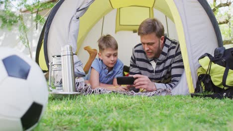 Caucasian-man-lying-in-a-tent-with-his-son-in-the-garden-using-a-smartphone