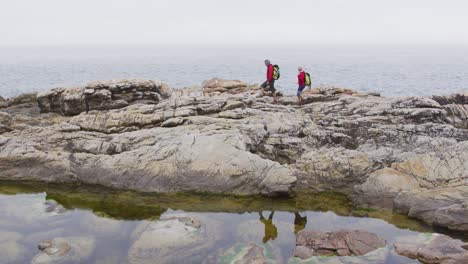 Senior-hiker-couple-with-backpacks-walking-on-the-rocks-while-hiking-near-sea-shore.
