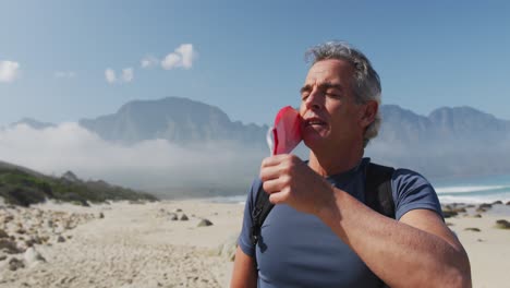 Senior-hiker-man-with-backpacks-removing-face-mask-and-breathing-while-hiking-on-the-beach.