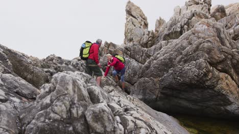 Senior-hiker-couple-with-backpacks-and-trekking-poles-holding-each-others-hands-while-climbing-rocks