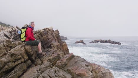 Senior-hiker-man-with-backpack-sitting-on-the-rocks-while-hiking-near-sea-shore.