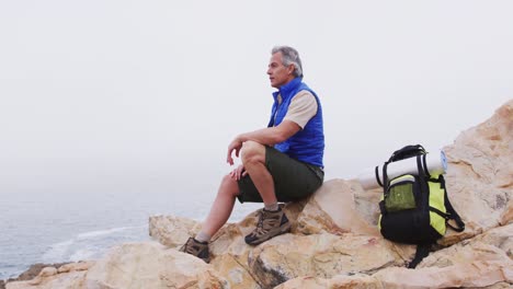 Senior-hiker-man-with-backpack-sitting-on-the-rocks-in-the-mountains-and-watching-the-sea-shore.