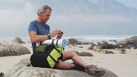 Senior-hiker-man-with-backpack-using-smartphone-while-sitting-on-rock-hiking-on-the-beach.