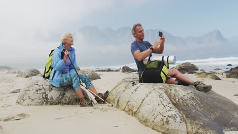 Senior-hiker-couple-with-backpack-and-hiking-poles-taking-a-selfie-from-smartphone