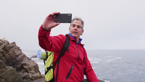 Senior-hiker-man-with-backpack-standing-on-the-rocks-and-taking-a-selfie-on-smartphone-while-hiking-