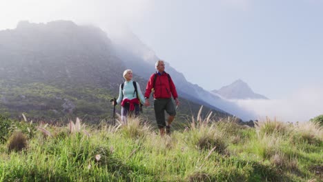 Senior-hiker-couple-with-backpacks-and-hiking-poles-holding-hands-and-walking-while-hiking