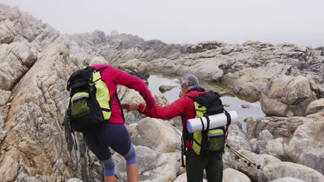 Senior-hiker-couple-with-backpacks-and-trekking-poles-holding-each-others-hands-while-climbing-rocks