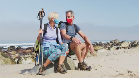 Älteres-Wanderpaar-Mit-Gesichtsmasken,-Rucksäcken-Und-Wanderstöcken-Redet-Auf-Einem-Felsen-Sitzend